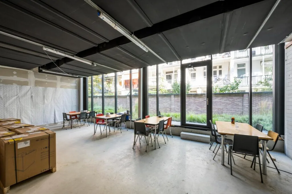 Gemeenschappelijke ruimte aan de Karel du Jardinstraat met tafels, stoelen en grote ramen met uitzicht op een binnentuin.